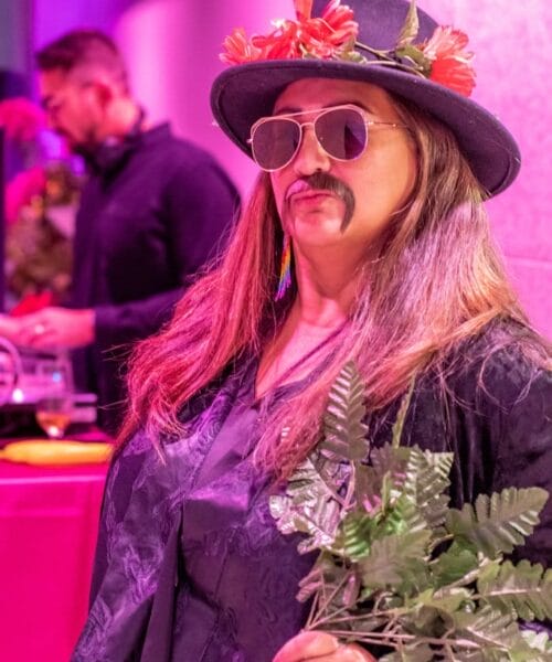 A person dressed in a black outfit with a hat adorned with flowers, a fake mustache, and sunglasses holds a bouquet of artificial greenery. In the background, another person can be seen standing near a table with various items under a pink and purple lighting.