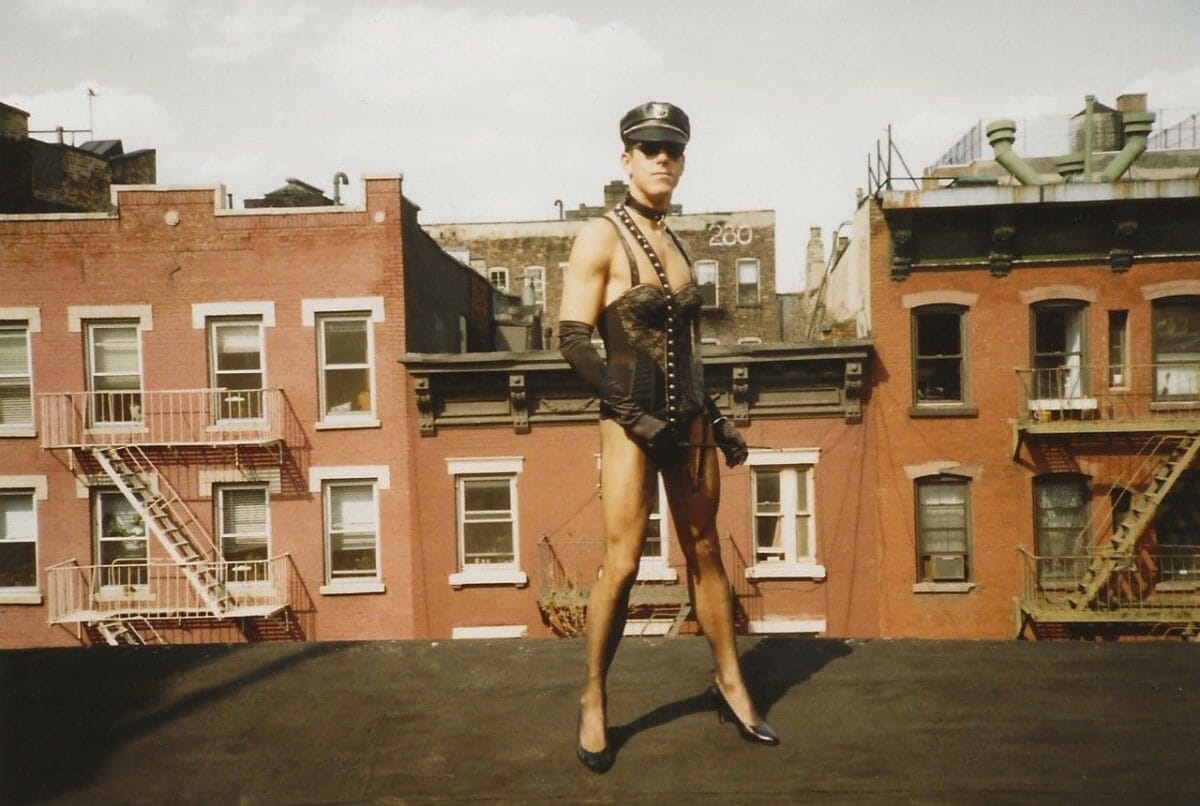 A person stands confidently on a rooftop wearing a police-style hat, sunglasses, long black gloves, a corset, high heels, and minimal clothing. The backdrop consists of red-brick buildings with fire escapes, set against a partly cloudy sky.