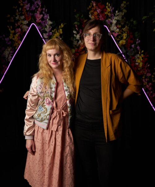 Two people stand close together in front of a dark background with neon pink triangular lights and colorful flowers. The person on the left wears a peach dress and embroidered jacket, and has long wavy golden hair. The person on the right has short dark hair and wears glasses, a black shirt, and an orange jacket.