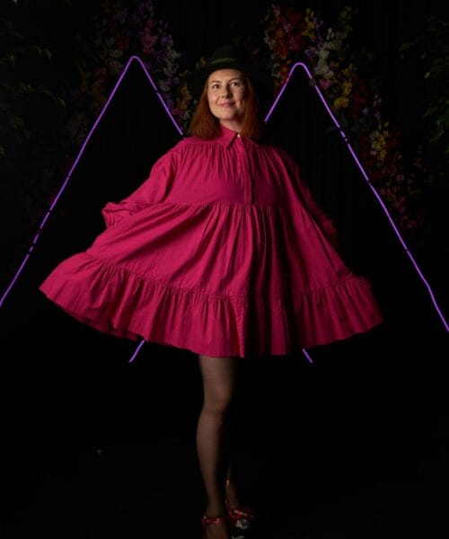 A person in a vibrant pink dress poses playfully, holding out the hem of the dress with both hands. They wear a hat and sparkling shoes, standing against a dark background with neon purple triangular shapes and colorful flowers.