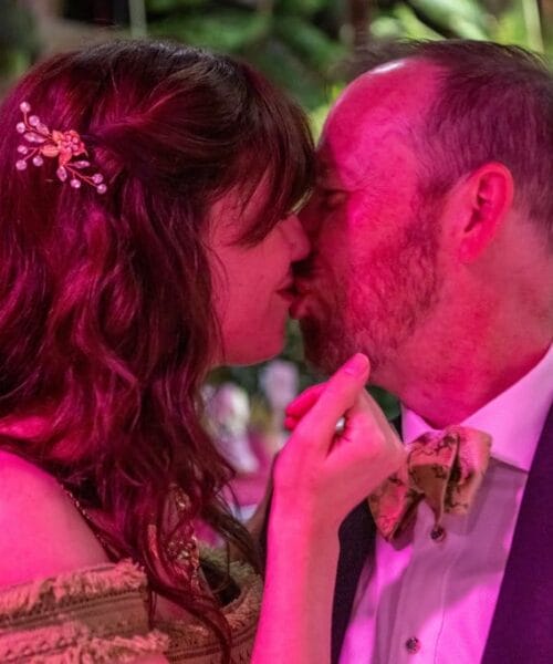 A couple shares a kiss in a warmly lit environment. The woman has long hair adorned with a decorative clip and wears an off-the-shoulder dress. The man has a beard and is dressed in formal attire with a bow tie. The background is softly blurred with hints of greenery.