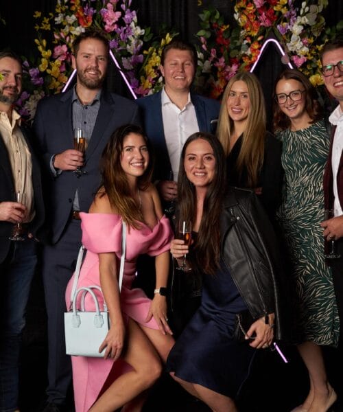 A group of eight people are posing together at what appears to be a formal event. They are standing in front of a backdrop with leaves and colorful flowers. Everyone is dressed in semi-formal attire, holding drinks, and smiling at the camera.