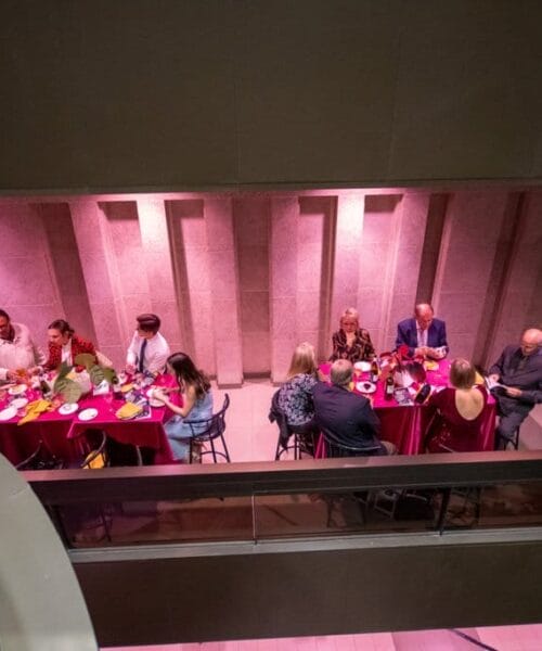 A view from above shows a group of people seated around a long, red-clothed table, engaging in conversation and dining. The setting is dimly lit with a purple hue, and the architecture features stone walls and a modern design.