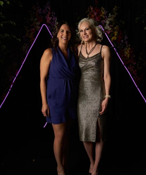Two women pose together in evening attire against a dark backdrop with illuminated triangular purple neon lights and floral decorations. One wears a blue sleeveless dress, while the other wears a silver, sparkling sleeveless dress with a side slit. Both are smiling.
