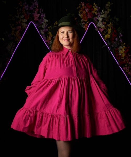 A woman wearing a pink dress and a green hat stands in front of a black background with neon purple mountain-shaped lights and colorful flowers. She holds the sides of her dress, creating a flowing effect. Trees frame the scene on both sides.