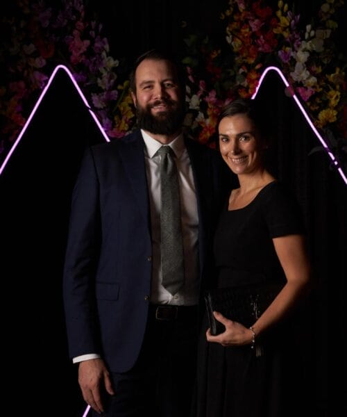 A man in a dark suit and tie stands next to a woman in a black dress. Both are smiling and standing in front of a backdrop featuring neon pink triangular lines and colorful floral decorations. The surroundings are dimly lit, giving a formal atmosphere.