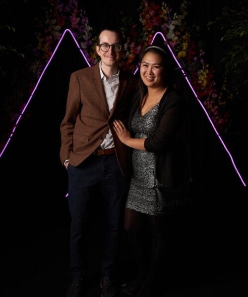 A man wearing glasses, a brown blazer, white shirt, and dark pants stands next to a woman in a sparkly silver dress and black jacket. They smile and pose together in front of a dark backdrop with purple neon triangle lights and floral decorations.