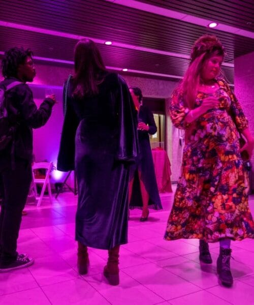 Four people are standing in a warmly lit room with a purple ambiance. One person on the right wears a vibrant floral dress, the others are engaged in conversation, and greenery is visible in the background on the left.