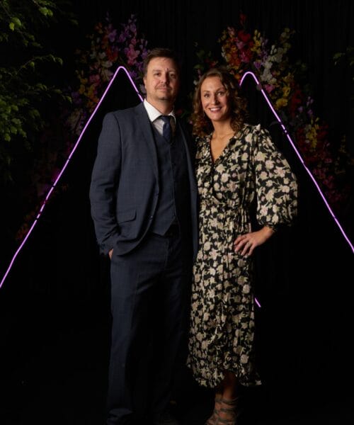 Two individuals, a man and a woman, are posing for a photo together. The man is wearing a blue suit, white shirt, and tie. The woman is wearing a patterned dress with floral designs. They stand in front of a dark backdrop with lit decorative elements and flowers.
