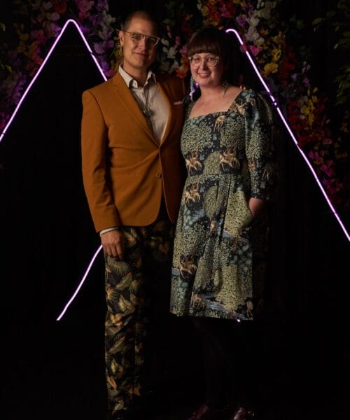 Two people are posing together against a vibrant and colorful backdrop. The person on the left is wearing a mustard jacket and patterned trousers, while the person on the right is wearing a patterned dress. Neon lights form triangles behind them, adding a modern touch.