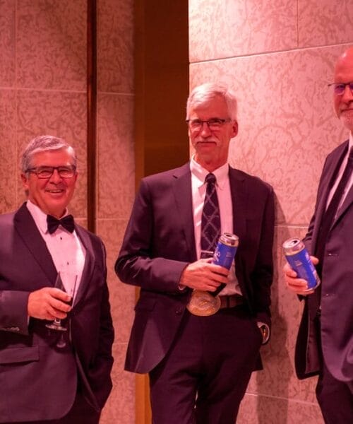 Three men in suits stand together at an event. The man on the left holds a glass of red wine, while the men in the middle and on the right hold cans of drinks. They are all smiling and appear to be enjoying their conversation. The background features patterned walls.