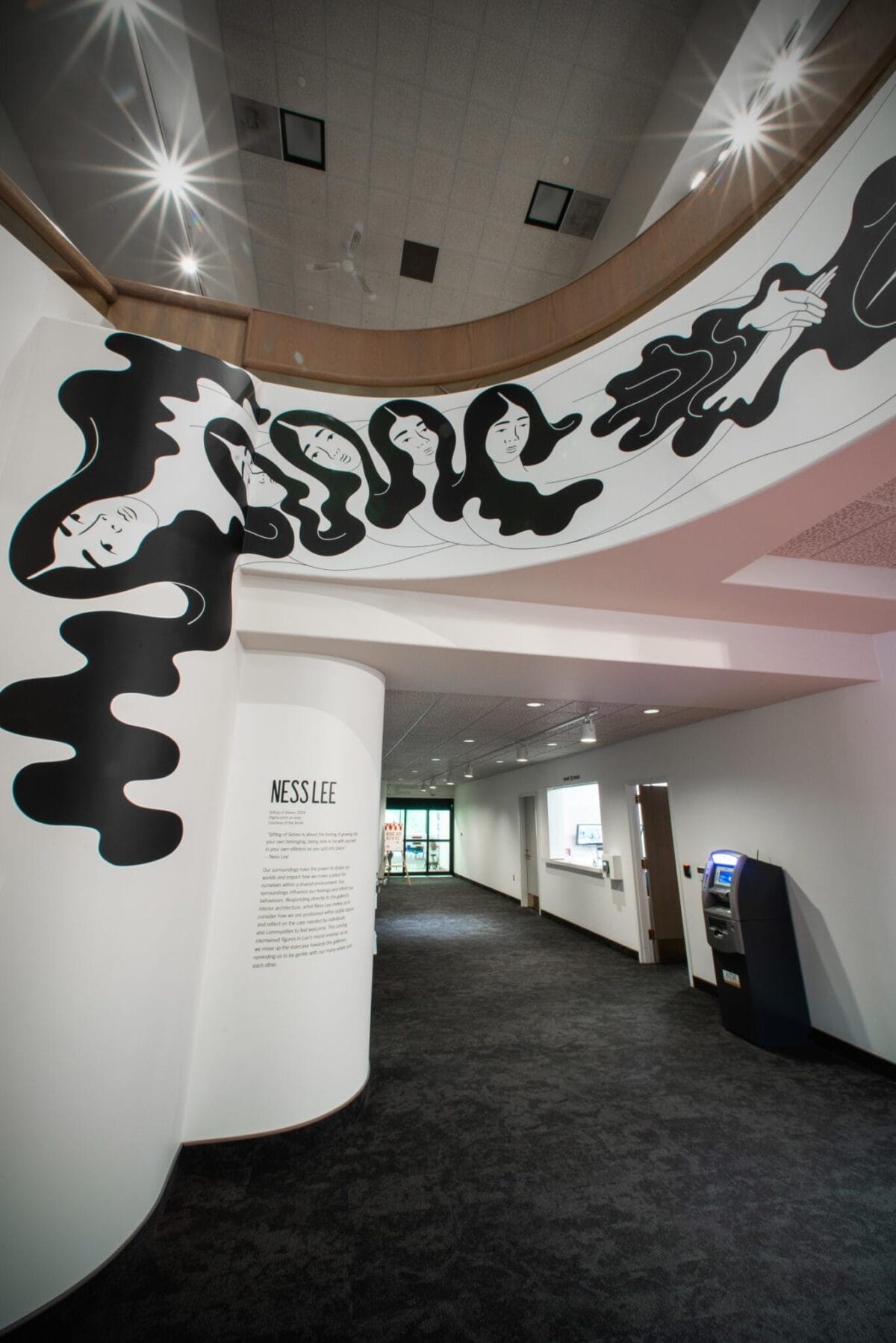 Large mural on a curved wall featuring abstract, flowing black shapes with faces. Below the mural, there is a description labeled 