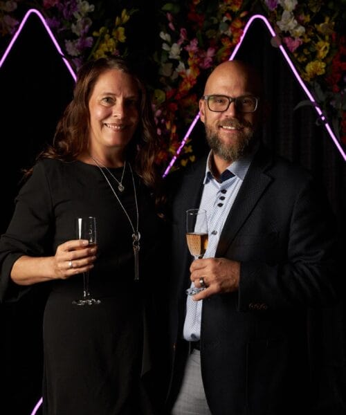 A smiling woman in a black dress and a man in a suit with a white shirt hold champagne flutes, posing together in front of a dark backdrop decorated with pink neon lights and colorful flowers.