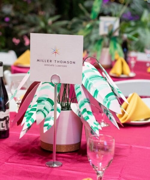 A table setting at an event features a Miller Thomson sign in a white stand. The table is covered with a red tablecloth and set with yellow napkins folded into points. Nearby are wine glasses, a bottle, and plant decorations. The background shows greenery and lights.