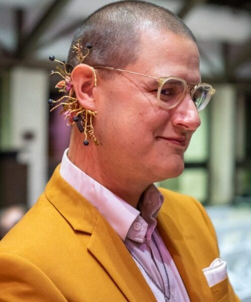 A person with short hair wearing glasses and a mustard yellow blazer over a light pink shirt. They have an intricate floral accessory tucked behind their ear, featuring small branches with delicate decorations. The background is blurred, showing an indoor setting.