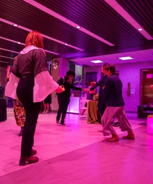 A group of people dance and socialize in a modern, brightly lit indoor space adorned with pink and purple lighting. Some people are gathered around a table in the background, while others dance in the foreground on the marble floor.