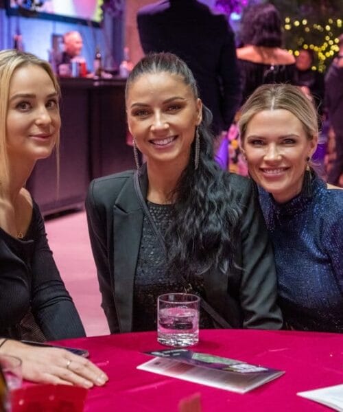 Three women smiling and sitting at a table during an evening event. They each have a drink in front of them: one has a glass of water, one has a glass of brown liquid, and one has a glass of white wine. The background shows other people socializing and festive lights.