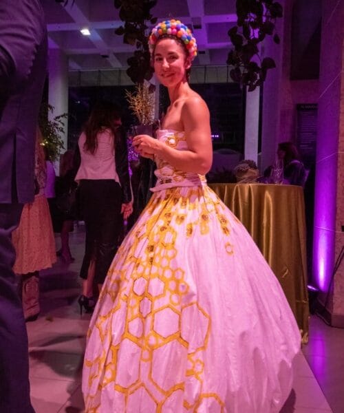 A woman stands in a dimly lit room at an event, wearing a white ball gown adorned with a bright yellow hexagonal pattern. She has a colorful pom-pom headband and her hair is styled neatly. In the background, other attendees are mingling near a table with a green cloth.