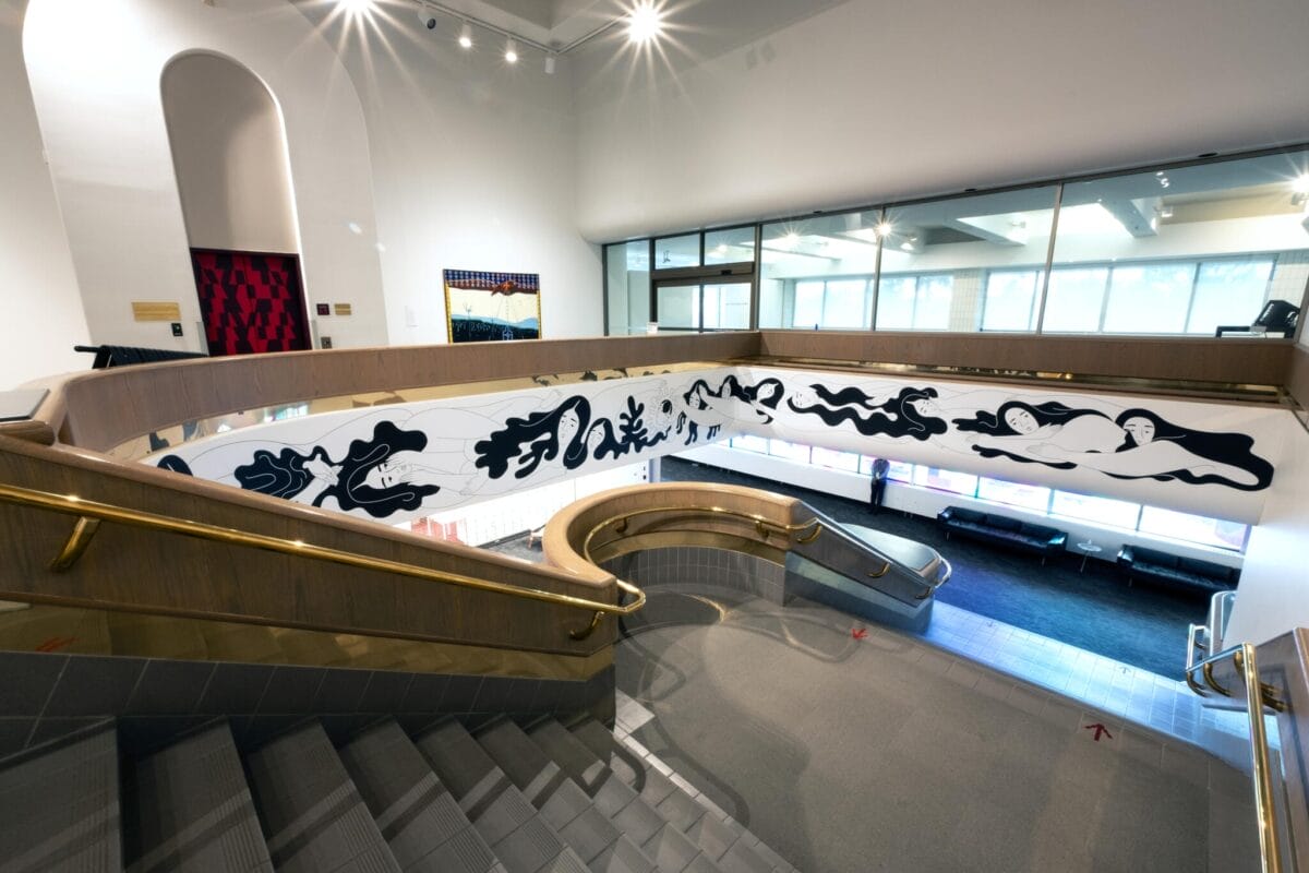 A modern interior features a curved staircase with a wooden railing. Below, a wall displays abstract black and white artwork. Large windows line the opposite side, allowing natural light to illuminate the space.