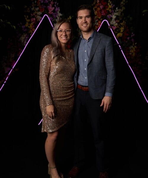 A woman in a sparkling gold dress and a man in a blue blazer and pants pose together in front of a black backdrop adorned with colorful flowers and pink neon light strips forming triangular shapes. They are both smiling and standing close to each other.