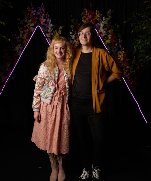 Two people pose in front of a black backdrop adorned with colorful flowers and two neon triangle outlines. The person on the left wears a pink dress, floral jacket, and boots, while the person on the right wears a mustard yellow cardigan, black shirt, and sneakers.