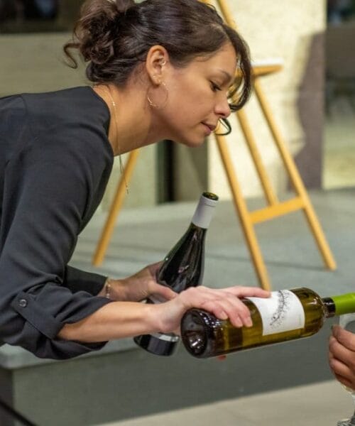 A woman with dark hair pours white wine into a wine glass held by someone off-screen. She holds a bottle of white wine in her right hand and has another bottle of wine tucked under her left arm. She is dressed in a black outfit and stands near an easel.