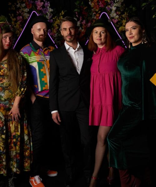 A group of five people stands posing in front of a dark background with colorful, triangular light decorations. They are dressed in vivid and eclectic outfits, with one person in a black suit, another in a pink dress, and others in creative, colorful attire.