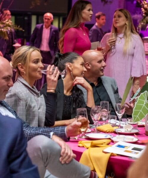 A group of people dressed in formal attire sit around a table at an event. They are engaged in conversation and laughter, holding drinks. The decor is elegant with pink lighting and floral arrangements in the background. A few people stand and chat in the background.
