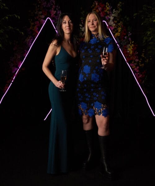 Two women dressed elegantly in evening attire pose with champagne glasses in hand. The woman on the left wears a long, teal gown, and the woman on the right wears a black and blue lace dress with black boots. A backdrop with floral decorations and purple neon lights frames them.