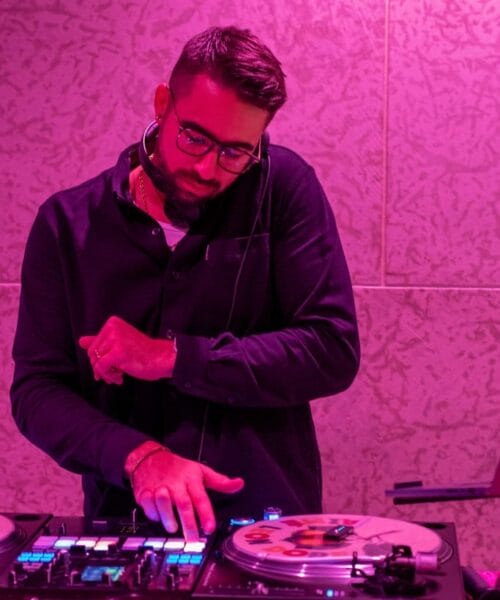 A DJ with short hair, glasses, and a beard is using a DJ console under pink lighting. He wears headphones around his neck, a black shirt, and appears focused on mixing music, with one hand on the turntable and the other adjusting controls.