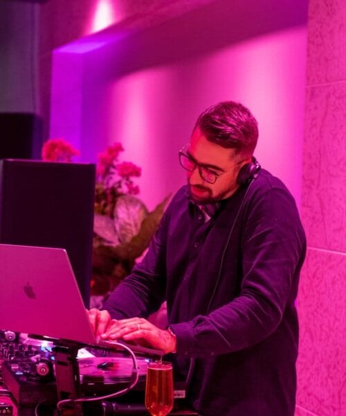 A man with short hair and glasses is DJing with a laptop and DJ equipment. He is wearing headphones and a dark long-sleeve shirt. The room is lit with purple lighting, and a glass of beer is placed on the table next to the DJ setup.