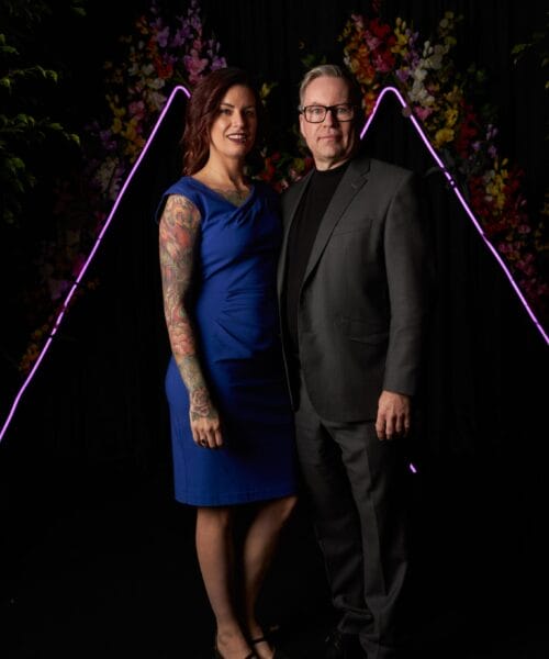 A woman in a blue dress and a man in a gray suit stand together in front of a dark backdrop with colorful flowers and pink neon lights shaped like triangles. The woman has colorful tattoos on her right arm. They are both smiling slightly.