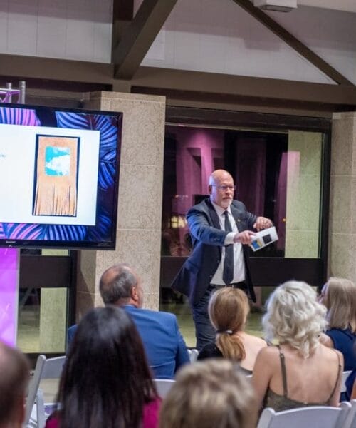 A man in a suit conducts an auction in a spacious room. Attendees sit on white chairs facing the auctioneer. A large screen beside the auctioneer displays 
