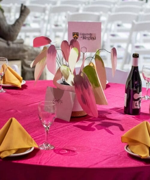 A round table with a bright pink tablecloth is set for a formal occasion. Each place setting includes a wine glass and a yellow napkin. The table centerpiece features paper decorations and a card with 
