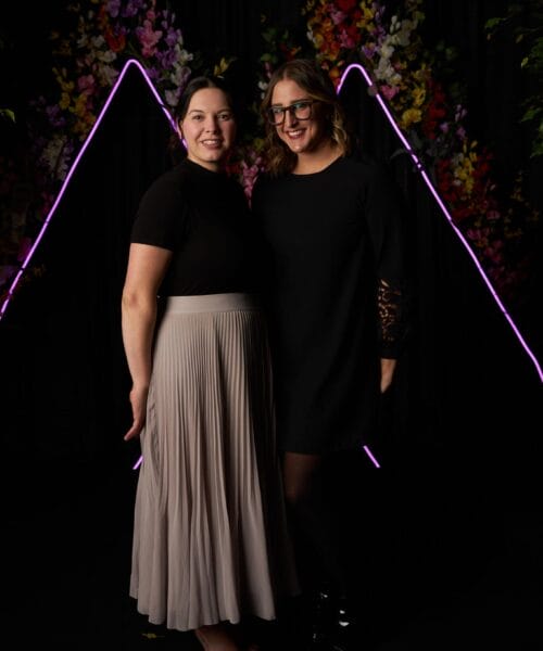 Two women are standing side by side in front of a dark backdrop adorned with vibrant, colorful flowers and neon triangle lights. One woman wears a black shirt and light beige pleated skirt, while the other is in a black dress with lace sleeves, smiling at the camera.