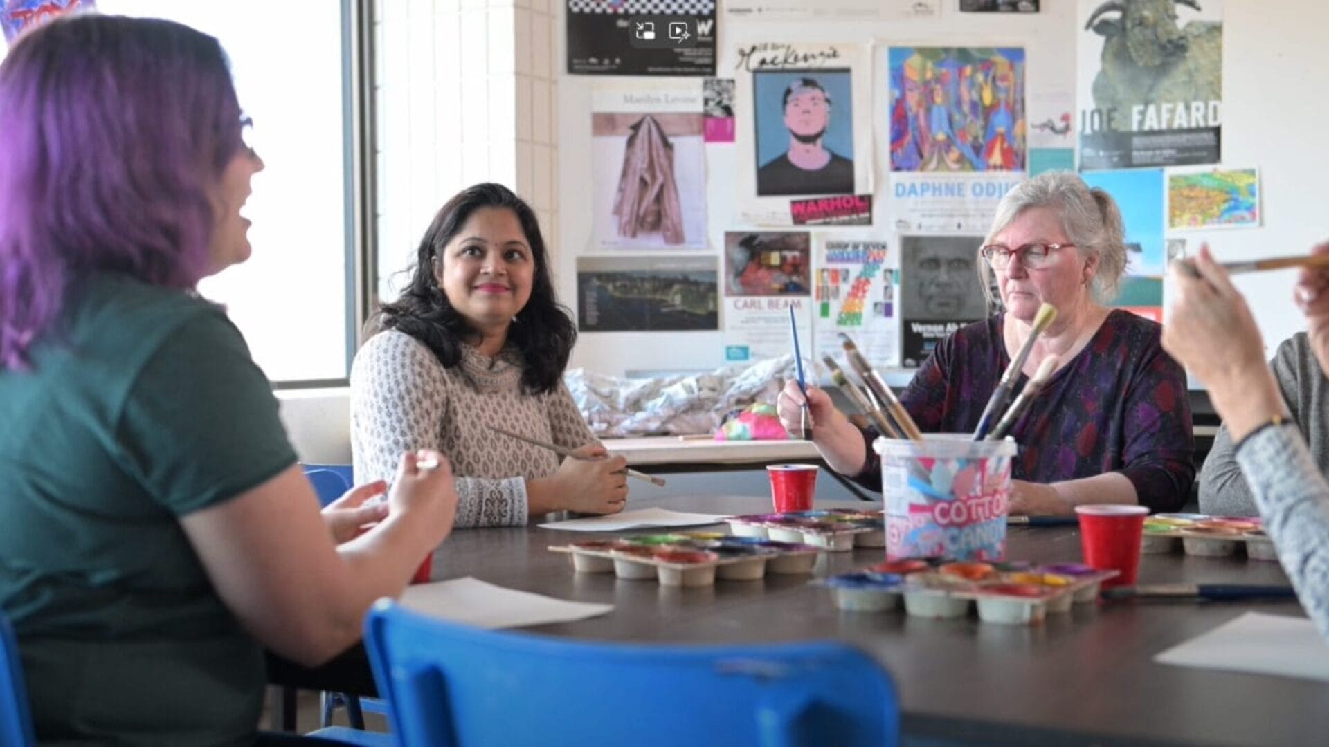 Four people are sitting around a table in a brightly lit room, engaging in a painting activity. Art supplies are scattered on the table, and colorful posters adorn the walls. The group appears relaxed and engaged in conversation.