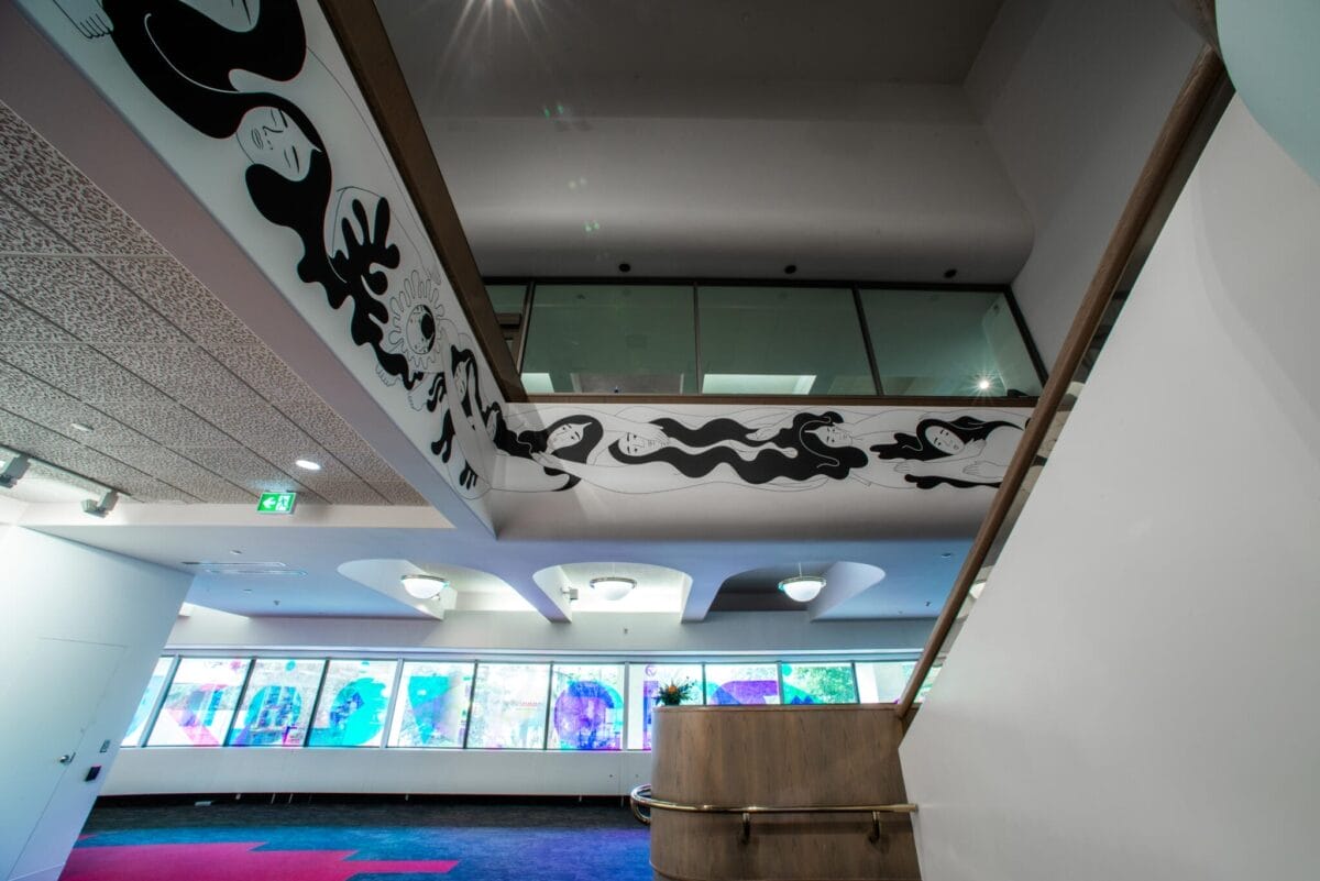 A modern interior features a stairway leading to an upper level with a glass balcony. A mural with abstract black-and-white illustrations decorates the wall. Large windows in the background display colorful, abstract art.