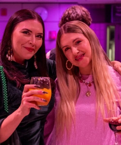 Two women are standing close together in a dimly lit room with a purple hue, smiling at the camera. Both are holding drinks; the woman on the left has an orange beverage, and the woman on the right has a glass of red wine. They appear to be at a social gathering.