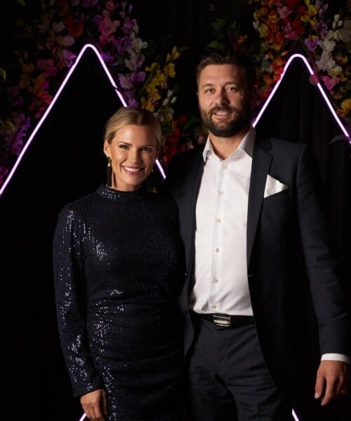 A smiling woman in a sparkly dark blue dress and a man in a black suit with a white shirt and pocket square stand together in front of an illuminated triangular backdrop with colorful flowers.