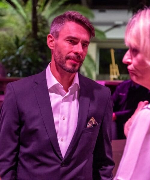 A man in a dark suit and white shirt converses with a woman in a white blouse at a social event. The background features people mingling and lush greenery under pink lighting. The setting appears to be a sophisticated indoor or outdoor venue.