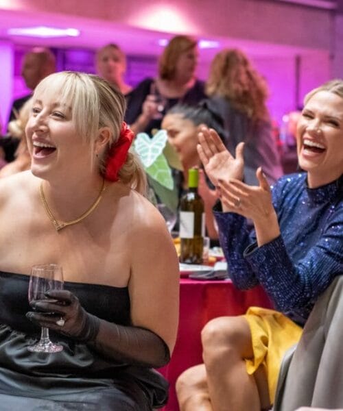 People at a lively event or party, smiling and laughing. A woman in a black dress holds a wine glass, another in a blue dress applauds. Both are seated at a table in a room with pink and purple lighting and other attendees in the background.