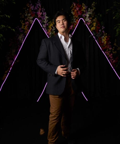 A person in a dark blazer and brown pants stands confidently in front of a backdrop featuring purple neon outlines of mountains and floral decorations. The individual has their hands clasped in front and looks directly at the camera.