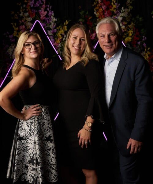 Three people are posing together in front of a dark background with colorful floral decorations and a triangular neon light arrangement. The two women are wearing black dresses, and the man is wearing a suit. They are smiling and standing close to each other.