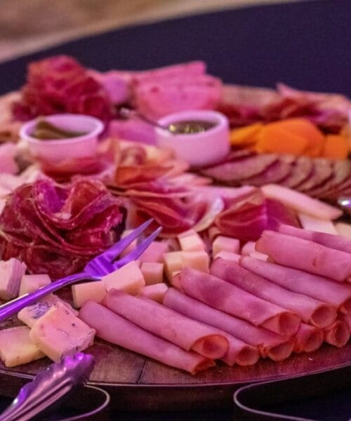 A charcuterie board with an assortment of cured meats, cheeses, and dipping sauces in small white bowls. The meats and cheeses are neatly arranged in rows and stacks on a round wooden board, with metal tongs placed on the side.