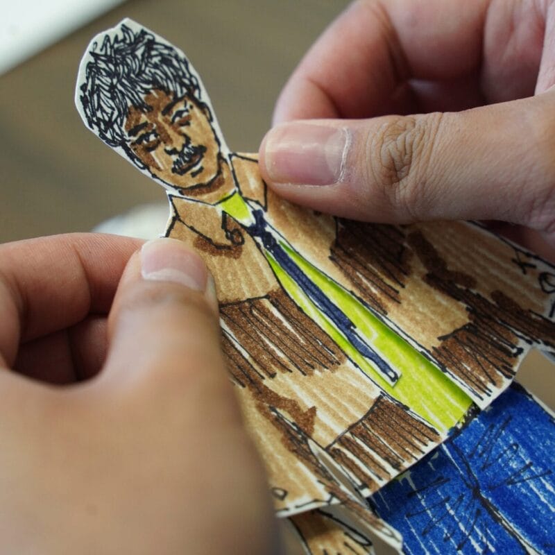 A person holds a hand-drawn paper cutouts doll of a man with curly hair, a mustache, a brown jacket, a green shirt, a blue tie, and blue pants. The paper doll is being carefully adjusted by two hands.