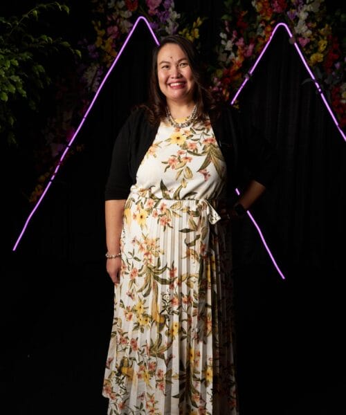 A woman smiles while standing in front of a dark backdrop with neon triangle lights and colorful floral decorations. She is wearing a white dress with a floral pattern, a black cardigan, and accessorized with a necklace and bracelet.