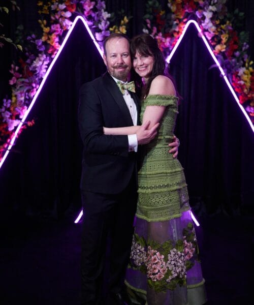 A man in a black tuxedo and bow tie and a woman in a green lace dress with floral details pose together, smiling, in front of a backdrop featuring purple neon lights and vibrant, colorful flowers.