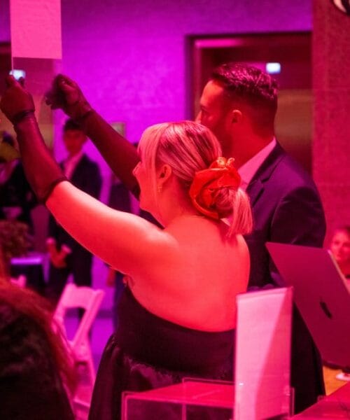 People in formal attire are enjoying an event in a pink-lit room. A woman with a flower in her hair is holding up a paper, possibly participating in an auction. A man in a dark suit and others observe. Various items and decor are visible on tables.