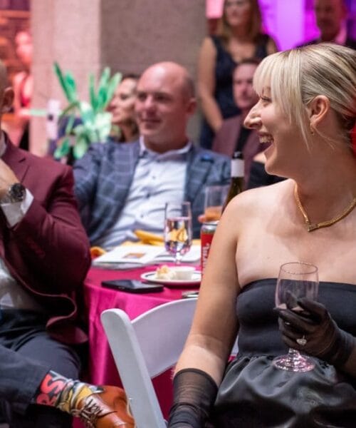 A group of people sit around a table at an event. A woman in the foreground, wearing a black strapless dress, long gloves, and a red flower in her hair, is laughing and holding a glass. Two men in suits are seated nearby, one wearing a red jacket and another in a blue jacket.