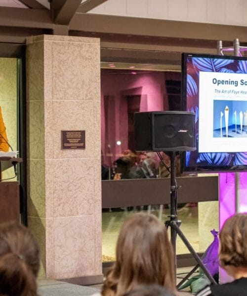 A speaker standing at a podium gestures while giving a presentation; a large screen to the right displays a slide titled 